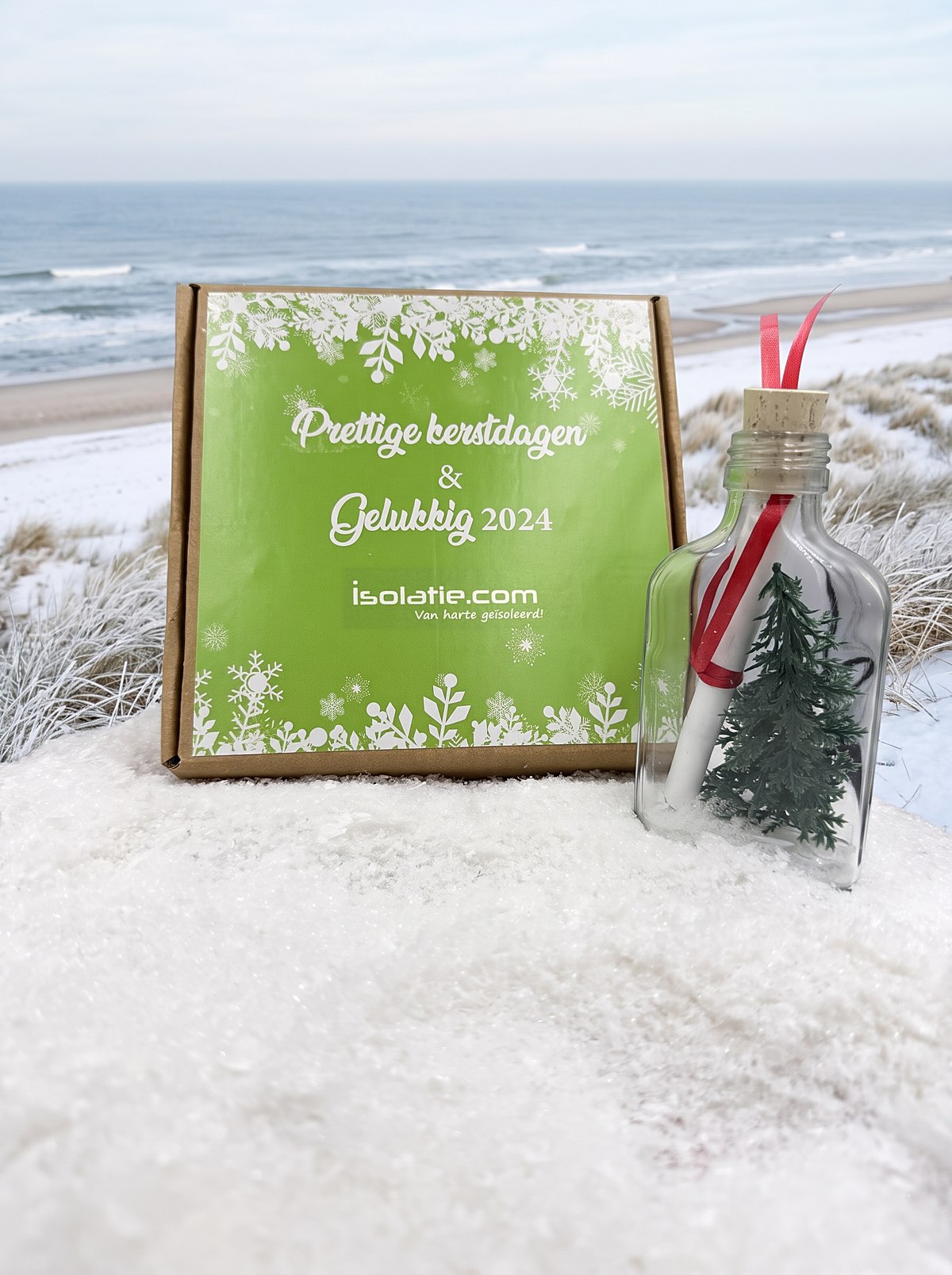 Kerstflessenpost voor isolatie.com: groen doosje met Prettige kerstdagen en Gelukkig 2024 en een flesje met opgerolde bon en kerstboompje, op een winterstrand aan zee