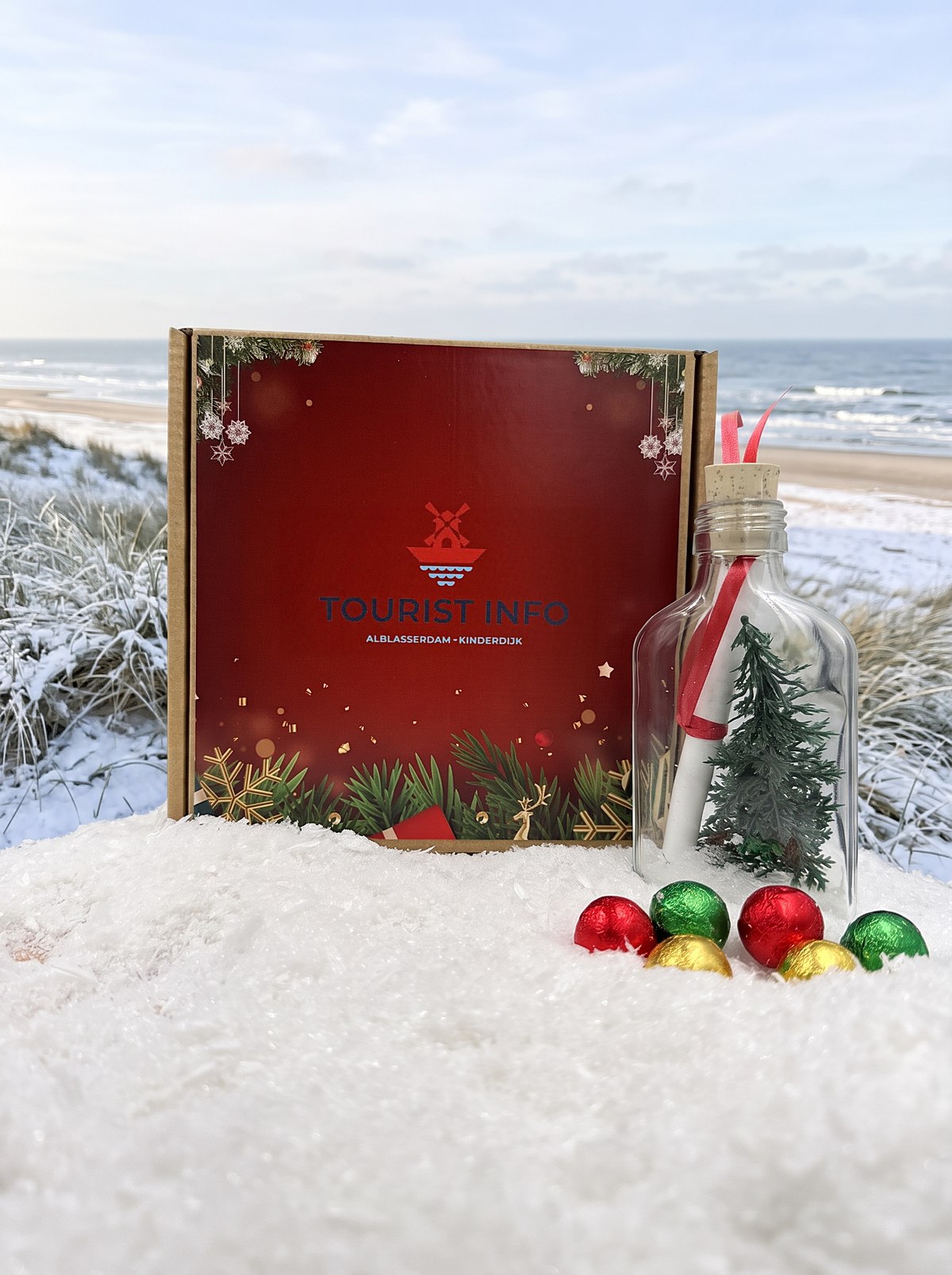 Nieuwjaarsflessenpost voor Tourist Info Alblasserdam-Kinderdijk: rood doosje met molenlogo, flesje met opgerolde bon en kerstboompje en chocoladekerstballen, op een winterstrand aan zee