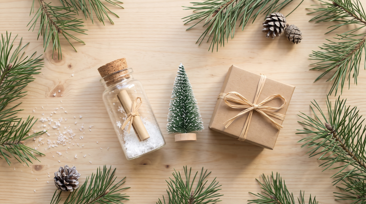 Glazen flesje met kurk, opgerolde boodschap en nepsneeuw naast een klein kerstboompje en een kraftpakje op een houten tafel