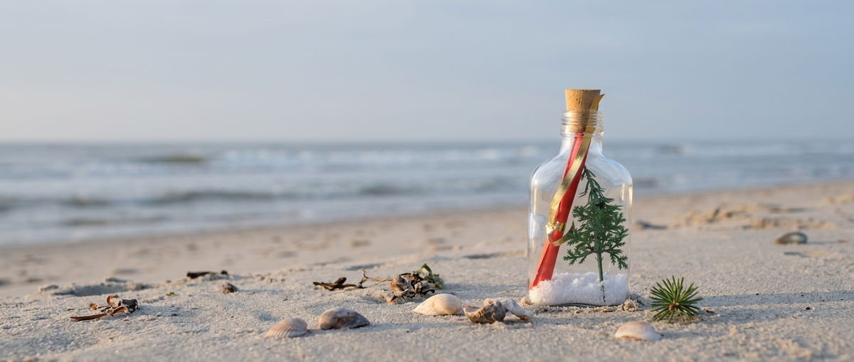 Kerstflessenpost: glazen flesje met nepsneeuw, kerstboompje en opgerolde uitnodiging in het zand aan zee